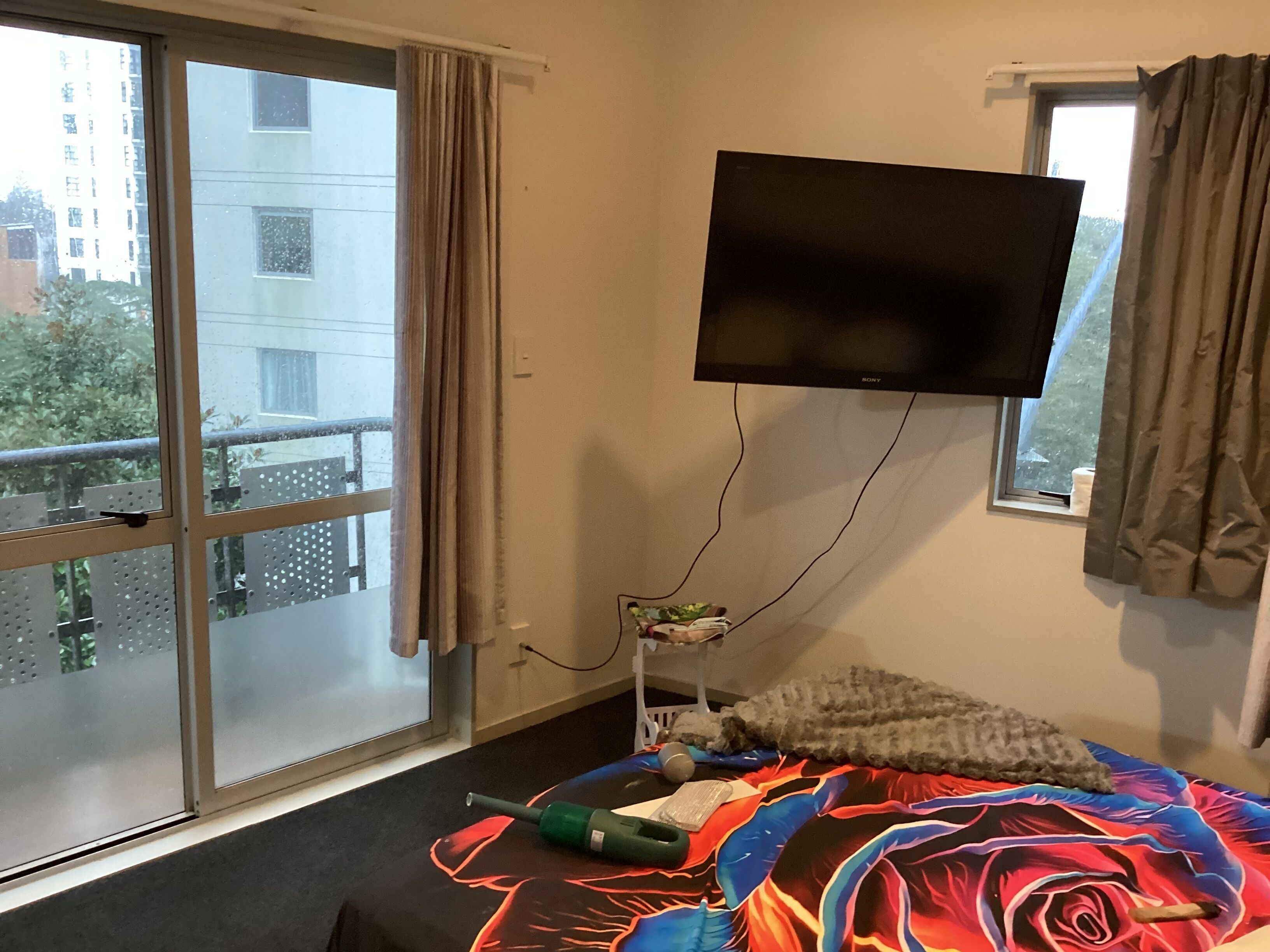 Een hotelkamer met een groot raam, een bed met een kleurrijke deken, een flatscreen televisie aan de muur en een klein nachtkastje met een lamp en een boek.