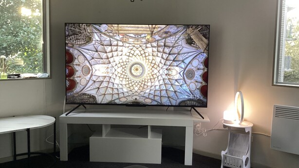A room with a large flat-screen TV mounted on a white stand, a small white side table, a white chair, and a wall clock. - 6a Scotia Place (Auckland)