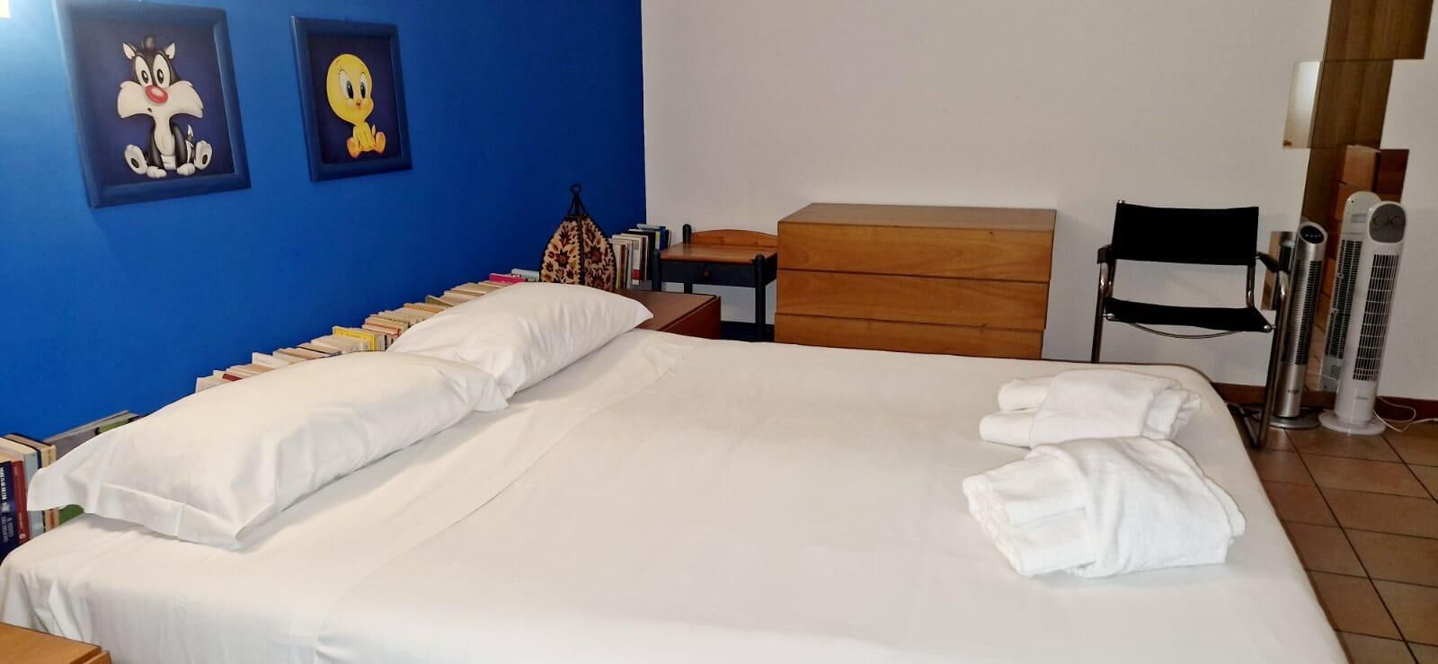 A neatly made bed with white linens, a wooden nightstand with a lamp, a chair, and a blue wall with cartoon character posters.
