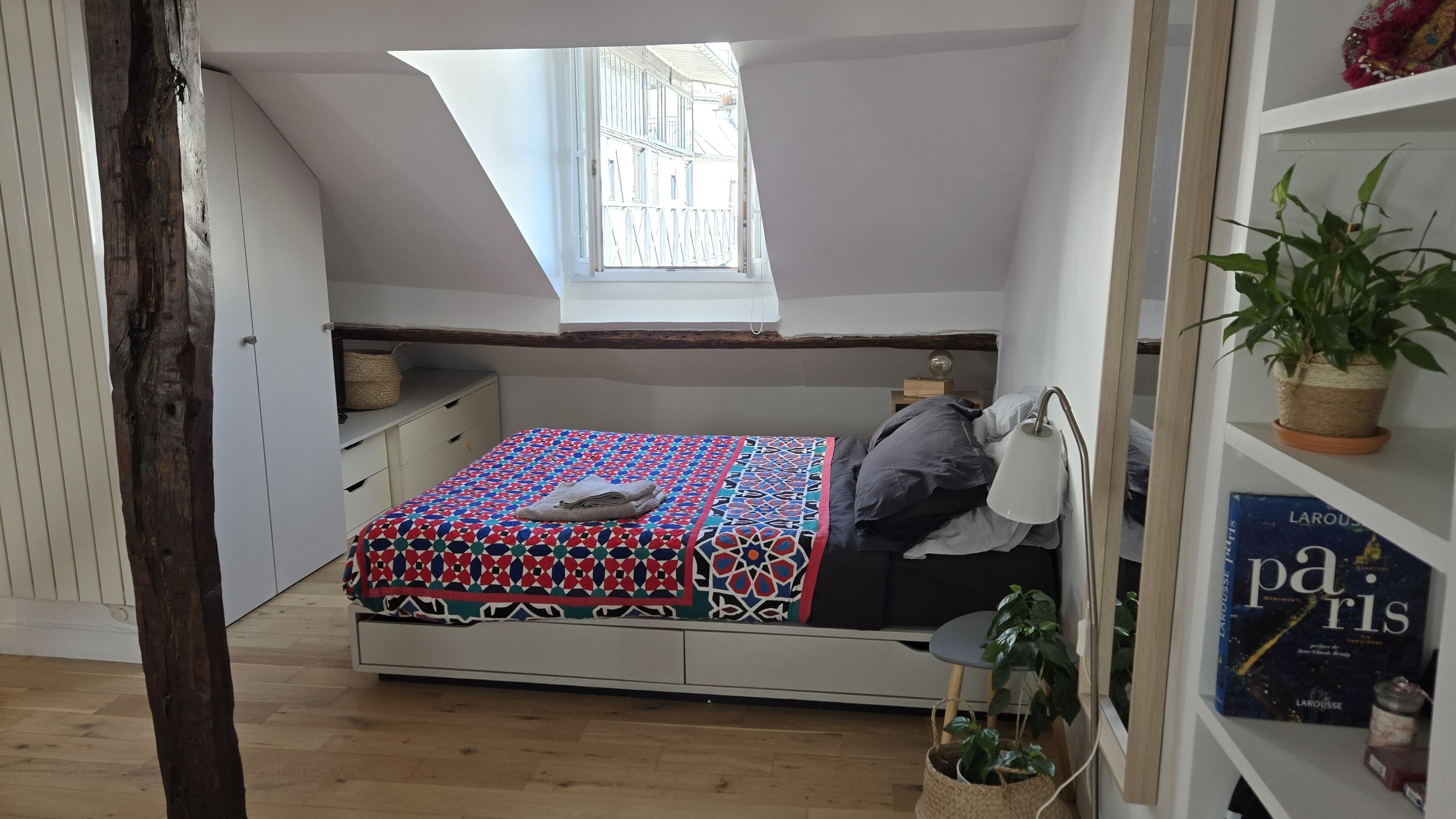 A bedroom with a sloped ceiling, a skylight, a bed with a patterned bedspread, a bedside table with a lamp, and a bookshelf with books.