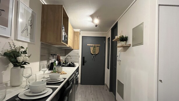 A narrow kitchen with wooden cabinets, a white countertop, and a dark door. There are framed pictures on the wall and a small plant on a shelf. - M&C Departamento República 815 (Santiago)