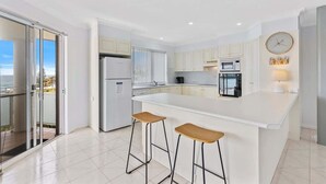 A modern kitchen with a central island, two bar stools, and a view through the glass door. - Sunrise on the Bay | Ocean Views & Beach (Malua Bay)