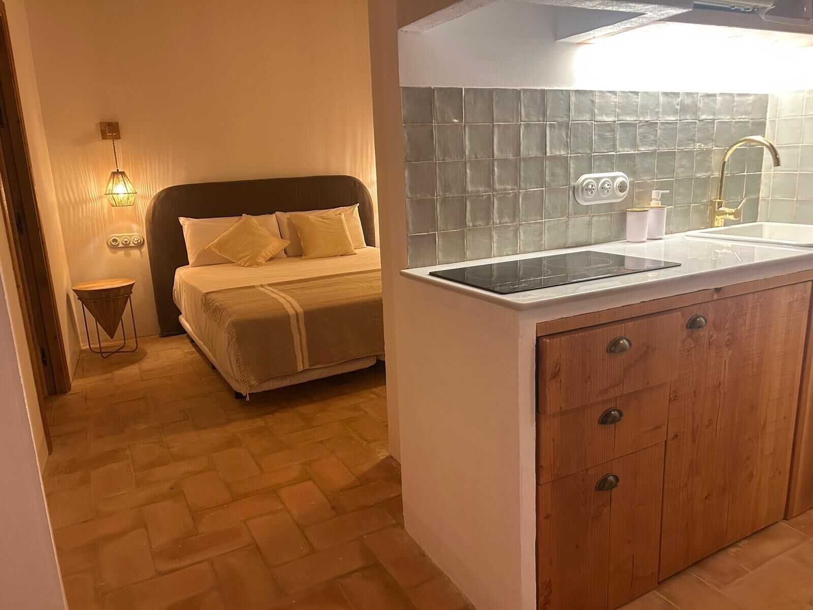 A bedroom with a bed, bedside table, and a small stool. There is a kitchenette with a sink, stove, and cabinets.