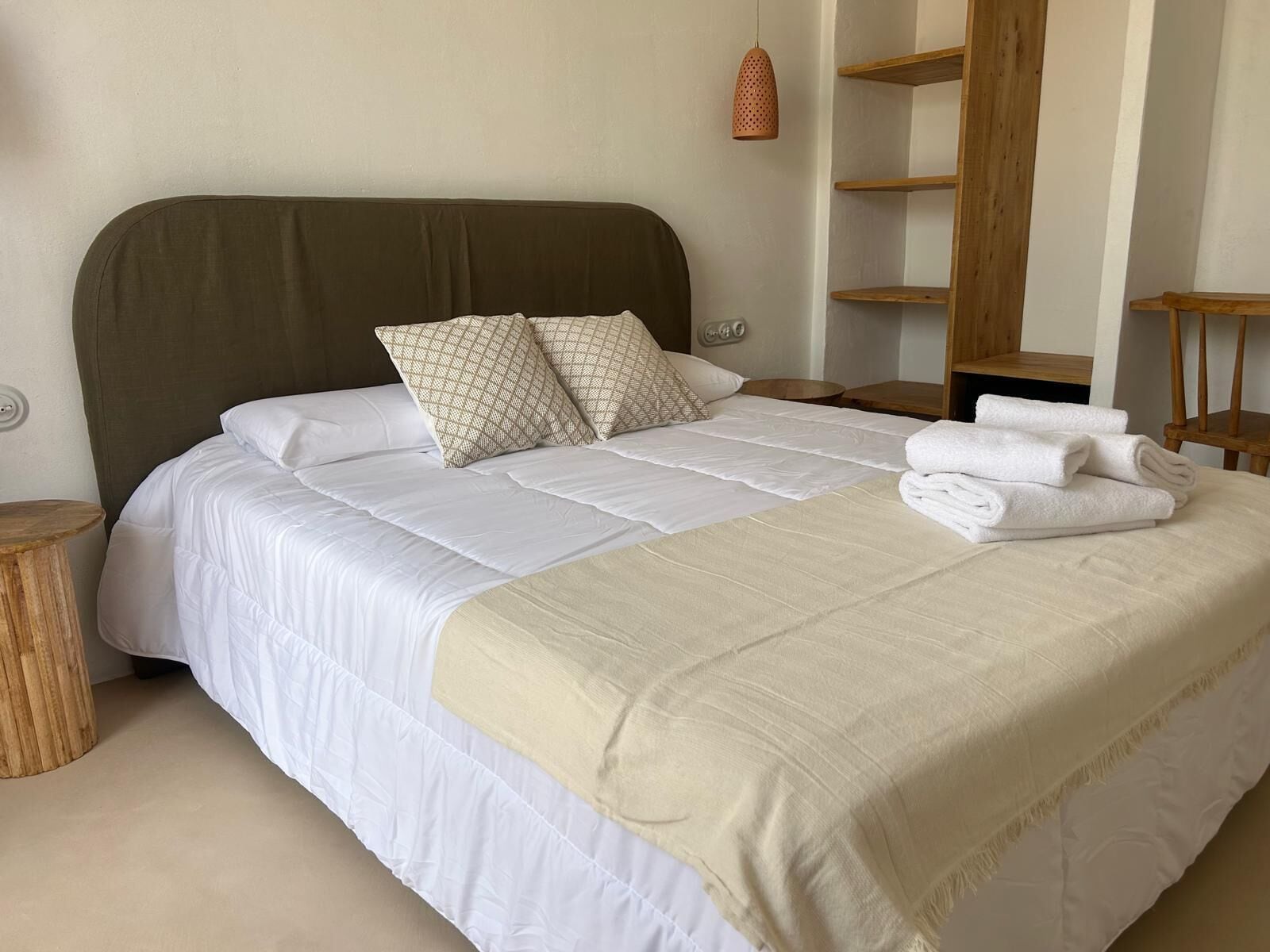 Ein ordentlich bezogenes Bett mit weißer Bettwäsche, zwei gemusterten Kissen und einer beigen Tagesdecke. Auf dem Bett liegt ein Stapel weißer Handtücher.