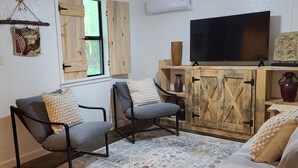 A cozy living room with two armchairs, a sofa, a wooden cabinet with a TV, and a window with wooden shutters.
