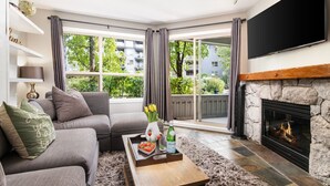 A living room with a grey sofa, a coffee table with a tray of fruit, a fireplace, and a large window with curtains.