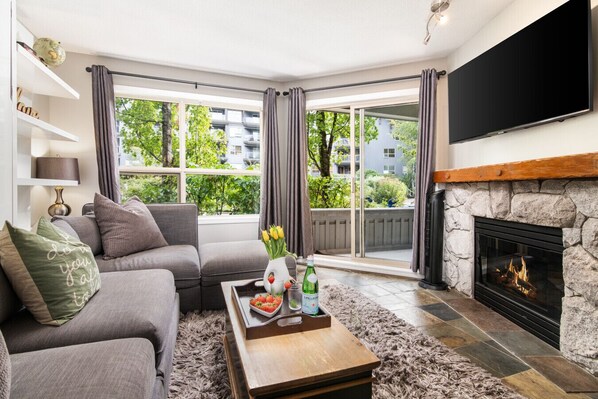A living room with a grey sofa, a coffee table with a tray of fruit, a fireplace, and a large window with curtains. - Ski-In/Ski-Out | On the Mountain! w/ Pool & Hot Tubs (Whistler)
