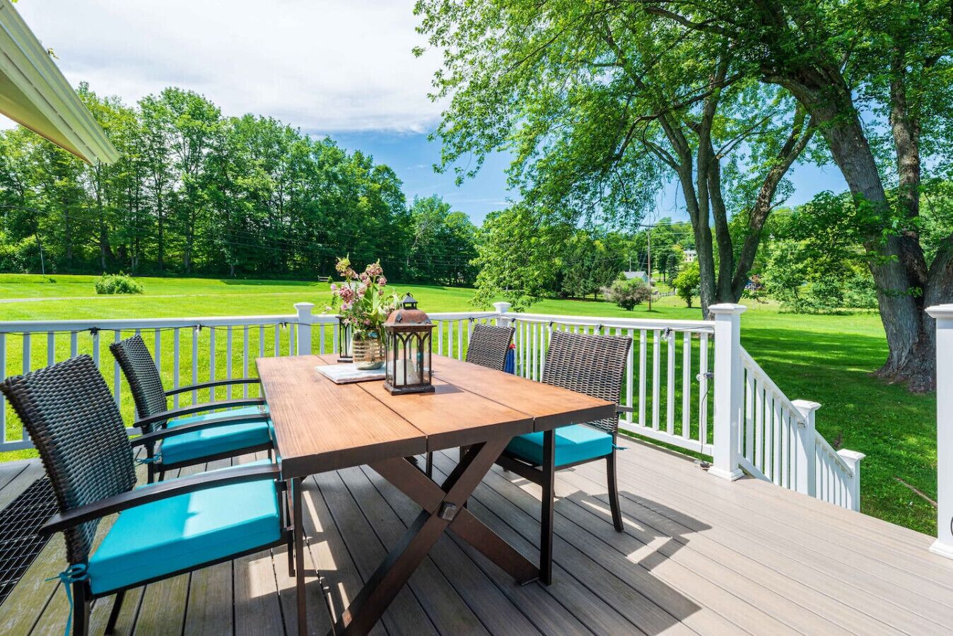 A wooden deck with a table and chairs, surrounded by white railings and a view of a green lawn and trees.