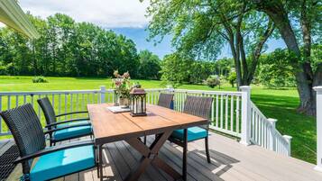 A wooden deck with a table and chairs, surrounded by white railings and a view of a green lawn and trees.