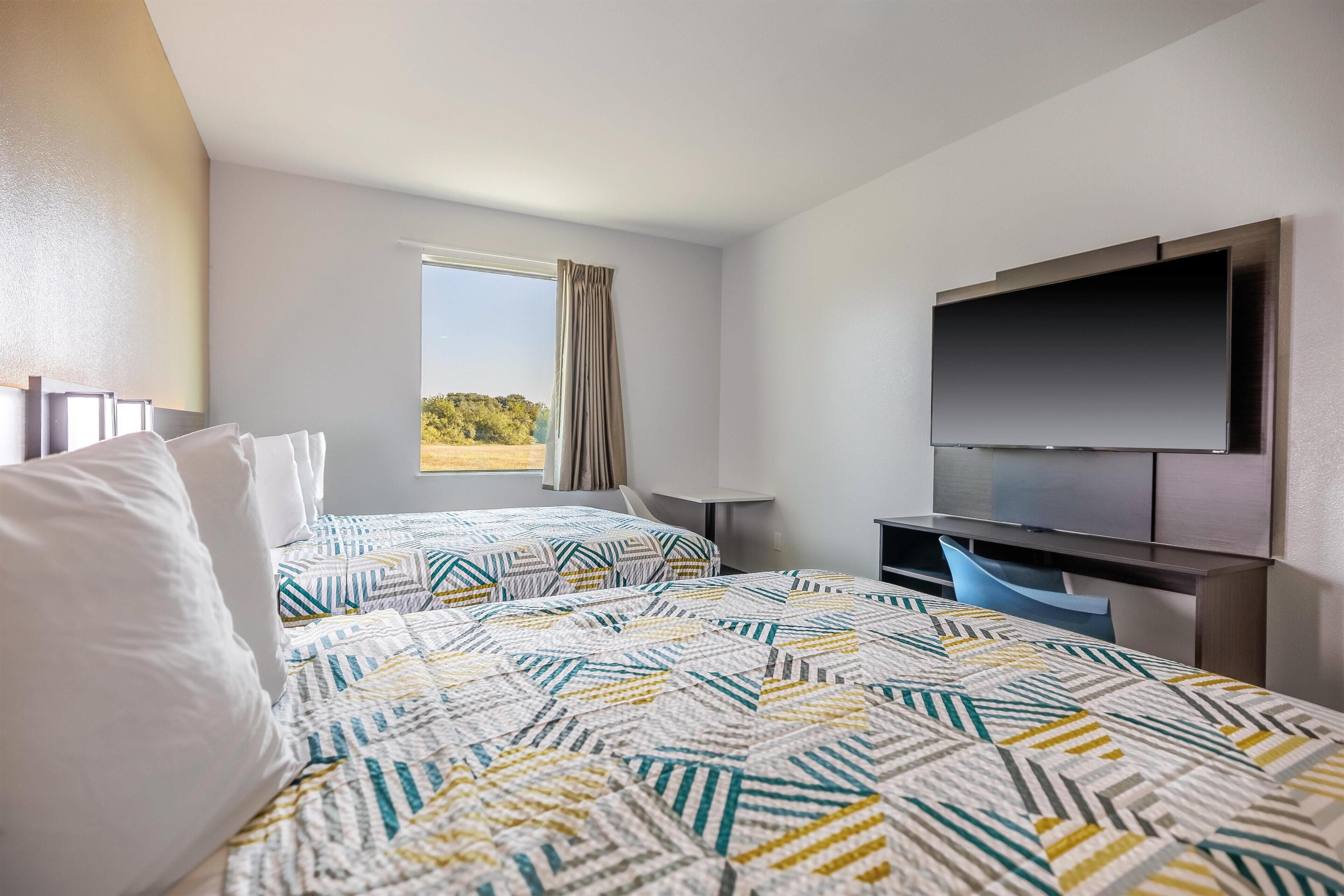 A hotel room with two beds, a TV, a desk, and a window with a view.
