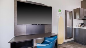 Une chambre d’hôtel moderne dotée d’un grand téléviseur à écran plat fixé au mur, d’un petit bureau avec une chaise, d’un miroir et d’un coin cuisine.