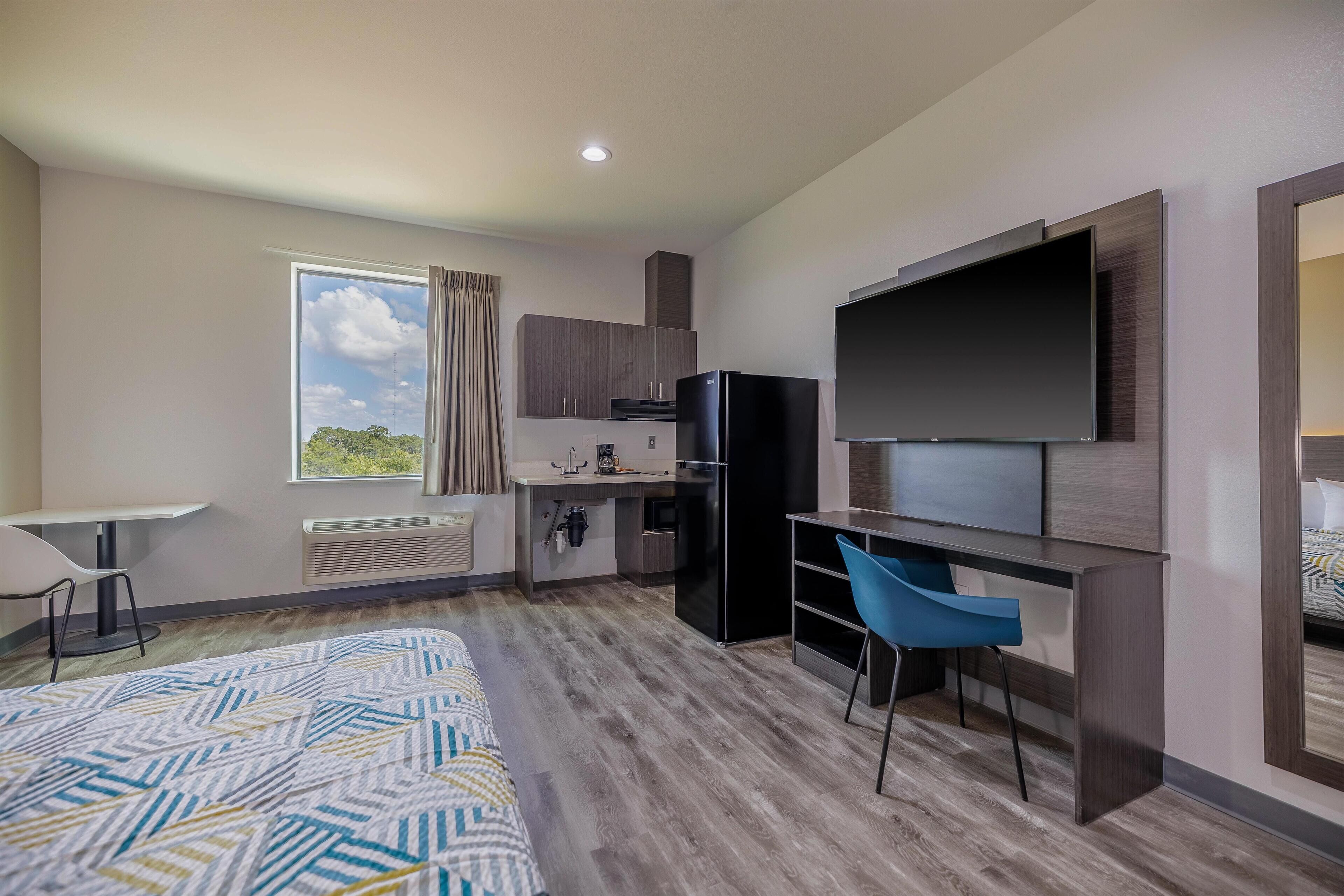 Una habitación de hotel moderna con una cama grande, un escritorio, una kitchenette y un televisor montado en la pared.