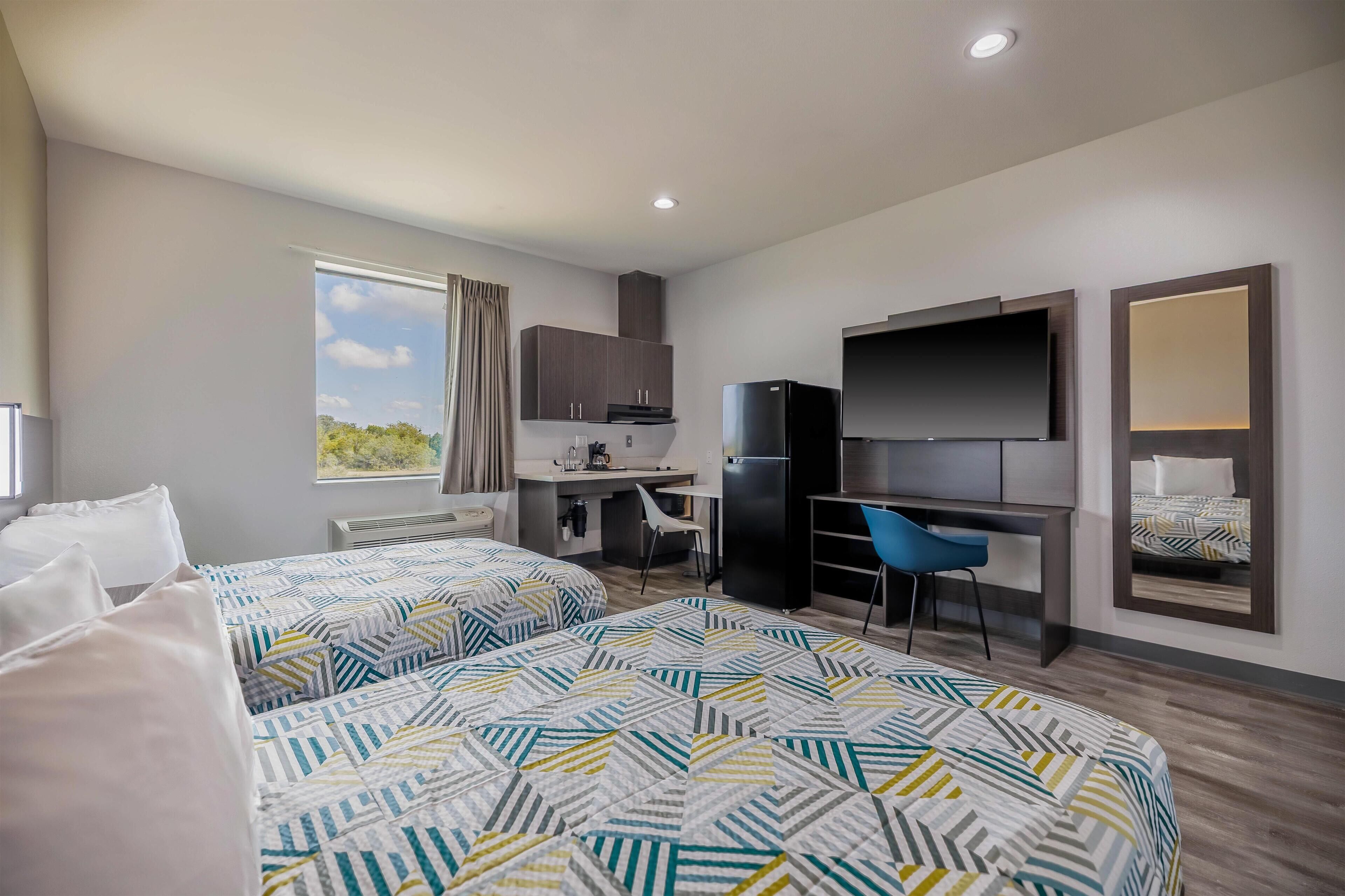 Una habitación de hotel moderna con dos camas, un escritorio, un televisor y una kitchenette.