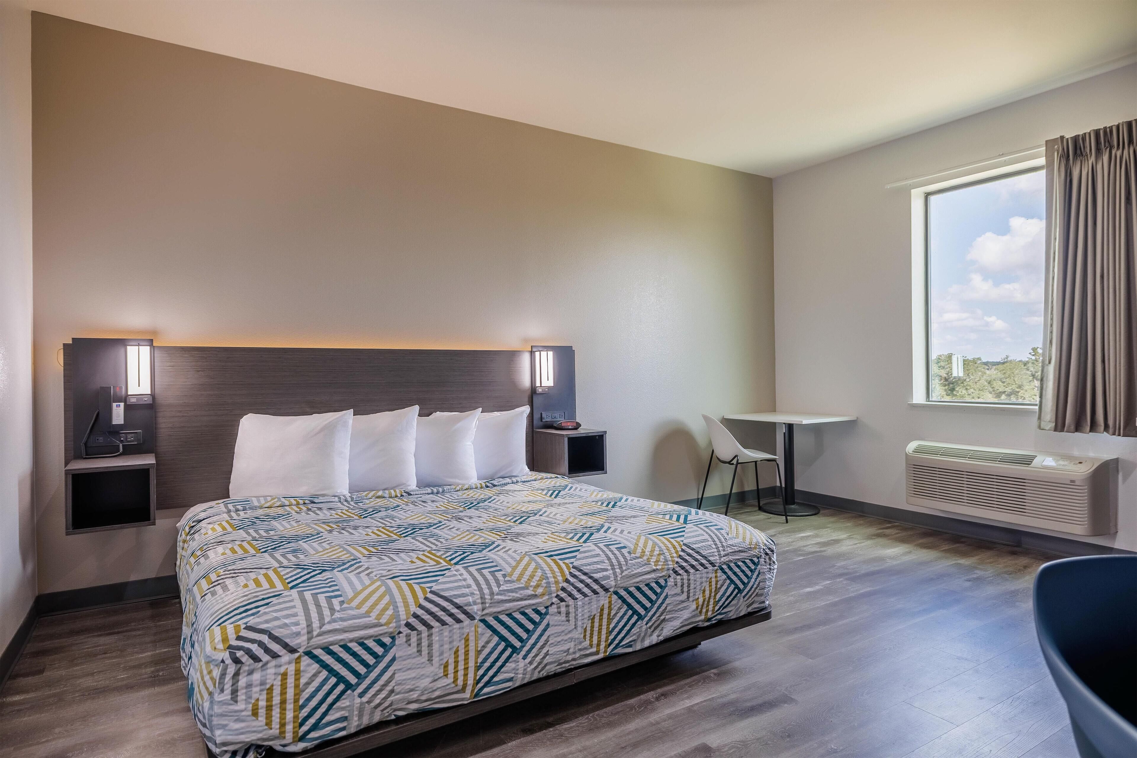 A hotel room with a large bed, bedside tables with lamps, a small table, a chair, and a window with curtains.