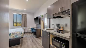 A compact hotel room with a kitchenette, a bed with patterned bedding, a blue chair, and a window with curtains.