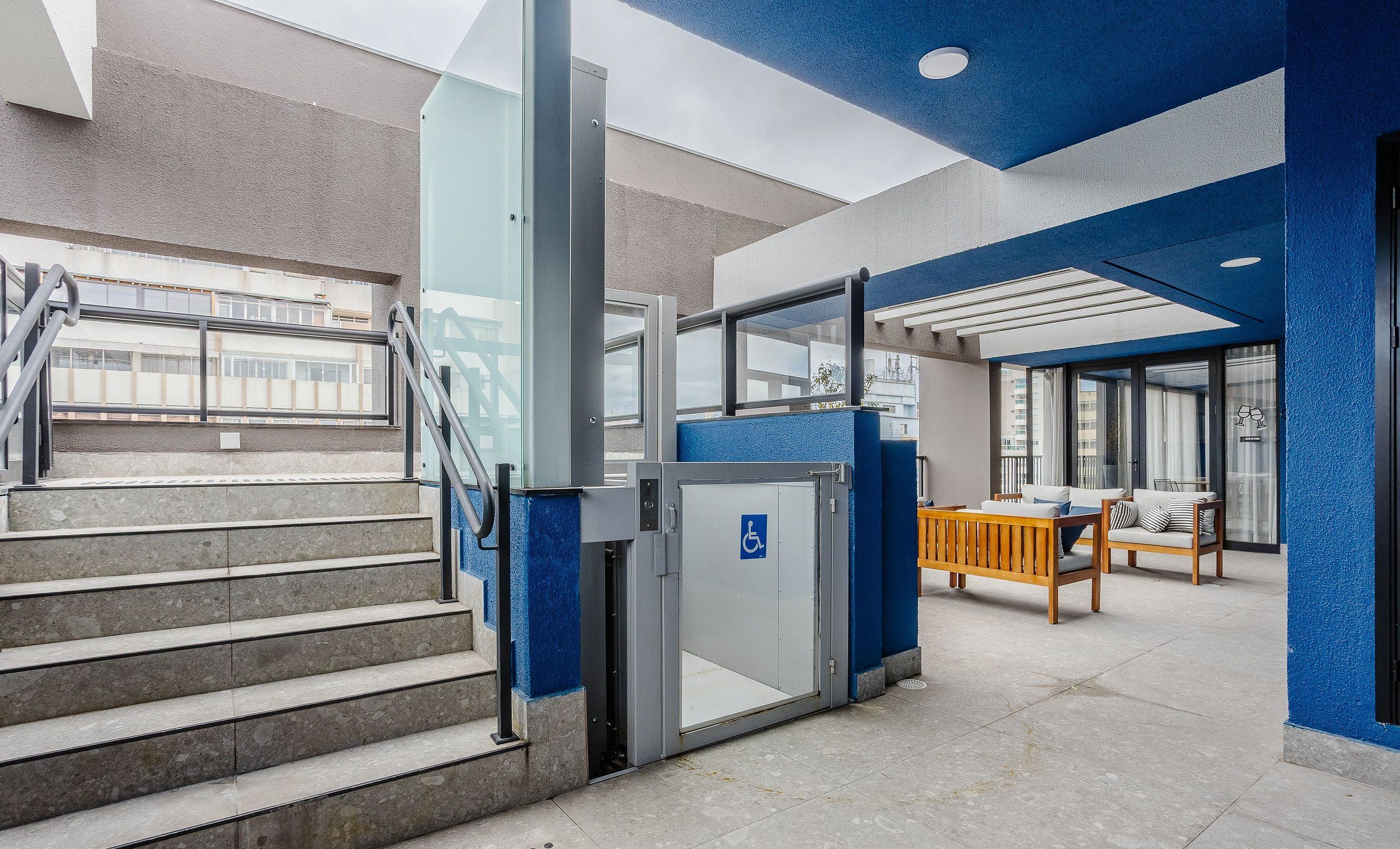 A modern building with a blue and grey color scheme, featuring a staircase, a wheelchair-accessible door, and a seating area with a table and chairs.