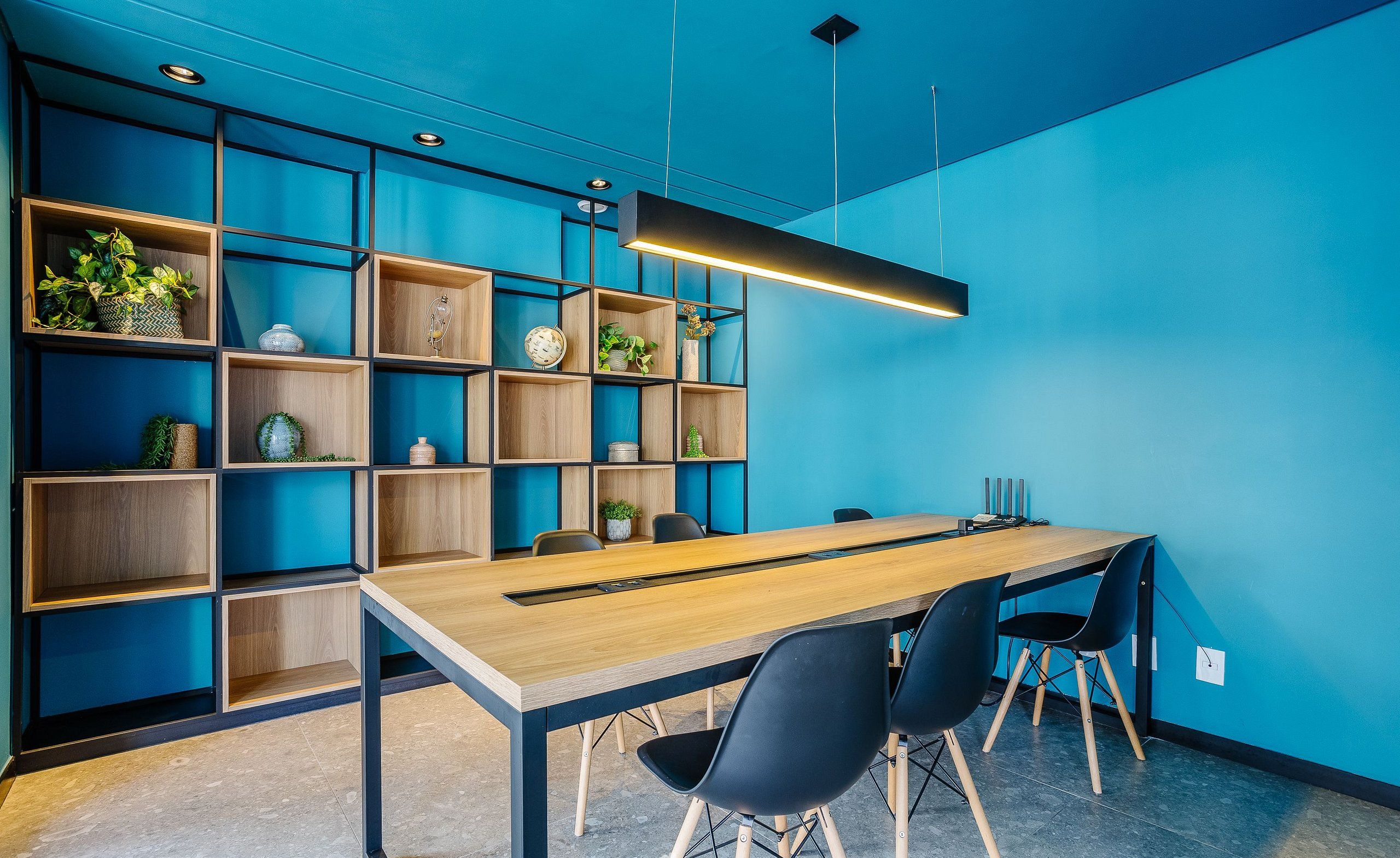 Uma sala de reunião moderna com uma longa mesa de madeira, cadeiras pretas e uma parede azul com estantes embutidas.
