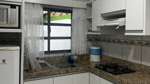 A compact kitchen with white cabinets, a built-in microwave, a gas stove, and a window with lace curtains. - Vista Bela Residential - Apt 07 (Centro Bombinhas)