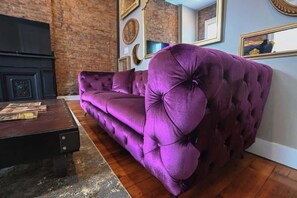A purple tufted sofa in a room with a brick wall and framed artwork. - NULU/Butchertown Key Club and Salon/Spa (Louisville)