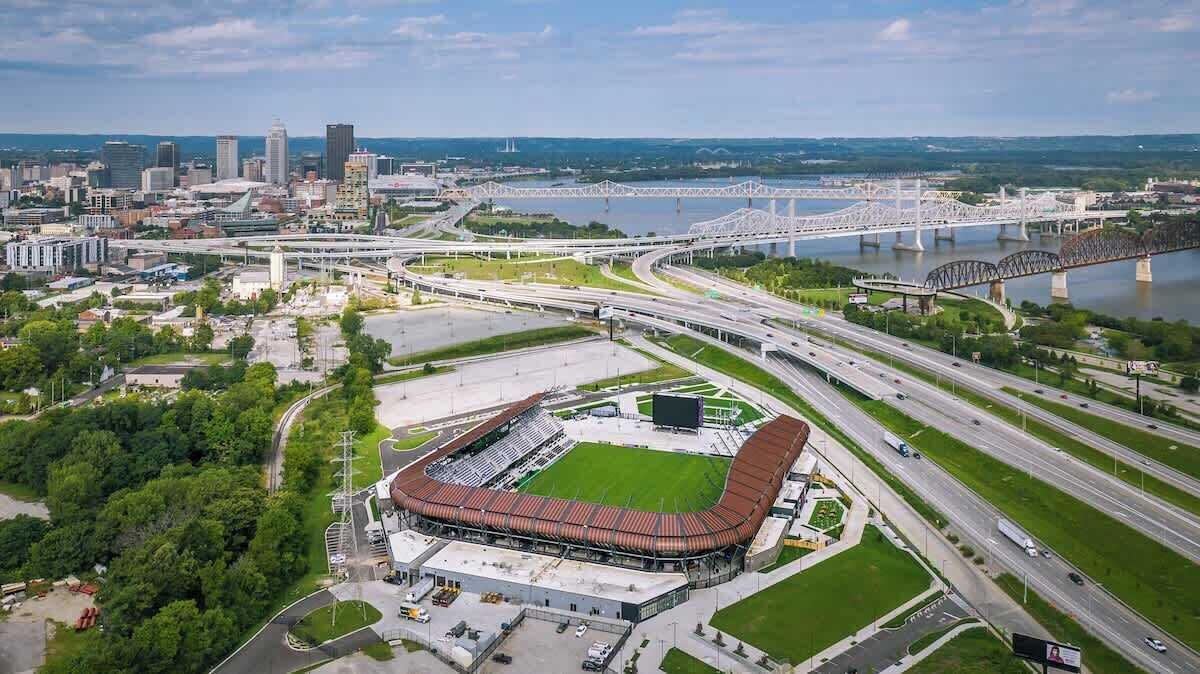 Une vue aérienne d’un stade avec un grand terrain, des routes qui l’entourent et un pont en arrière-plan.