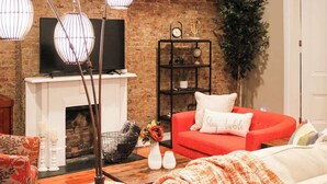 Una sala de estar con pared de ladrillo, chimenea, sofá rojo, sillón blanco y mesa de centro con un jarrón de flores.