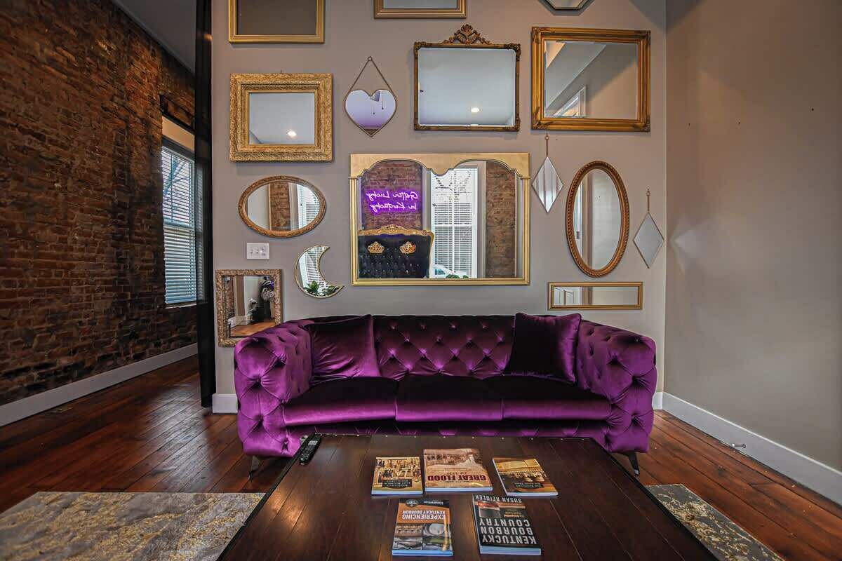 Une pièce avec un canapé en velours violet, une table basse sur laquelle sont posés des livres, et divers miroirs encadrés accrochés au mur.