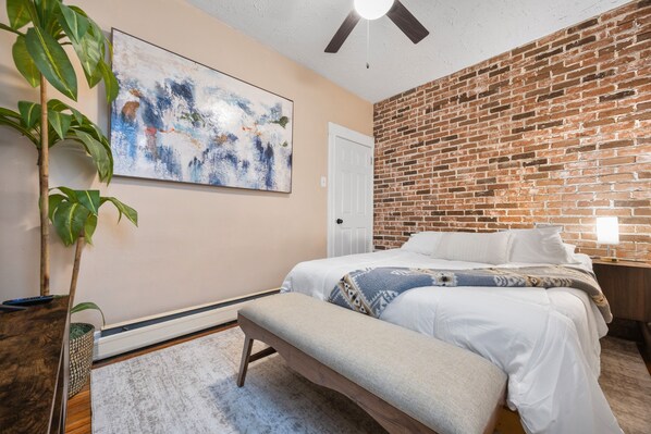 A bedroom with a brick wall, a bed with white bedding, a bench, a ceiling fan, and a large abstract painting.