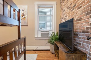 Une pièce avec un lit en bois, un téléviseur posé sur un meuble en bois, un mur en briques, une fenêtre avec des stores et une plante en pot.