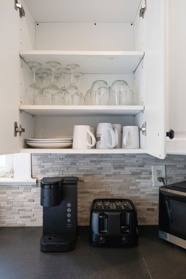 Una cocina con cafetera, tostadora y microondas. Hay estantes con cristalería y tazas blancas.