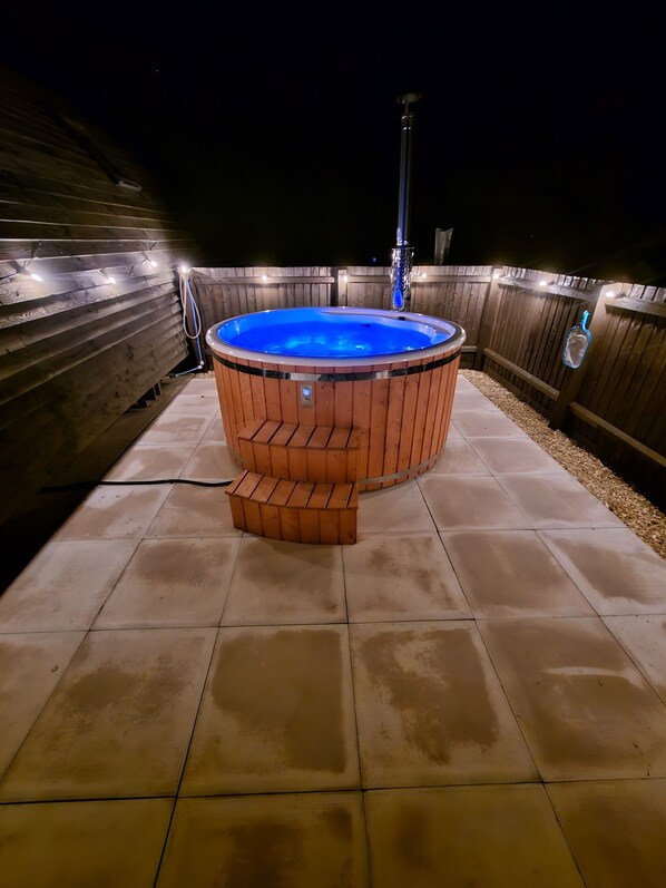 A hot tub with a wooden exterior and a blue interior, surrounded by a wooden deck and a tiled patio.