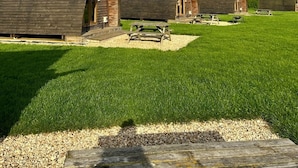 A row of rustic wooden cabins with pitched roofs, arranged in a grassy field with a clear sky above. - Cosy Rural Cabin with Wood Fired Hot Tub (Grantham)