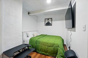 Une chambre avec un lit vert, une tête de lit blanche, une petite table, un banc, un téléviseur fixé au mur et un cadre photo.