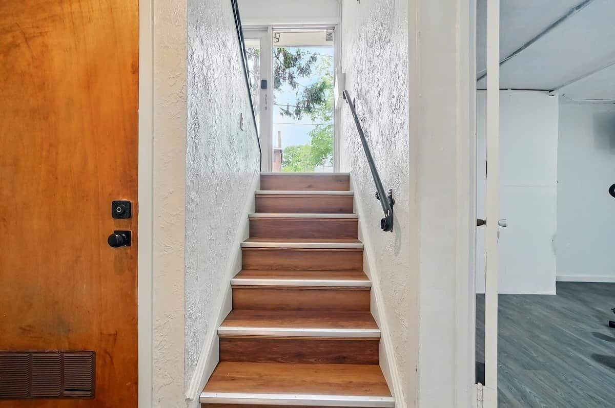 A wooden staircase leading to a door with a vent, white walls, and a view of greenery outside.