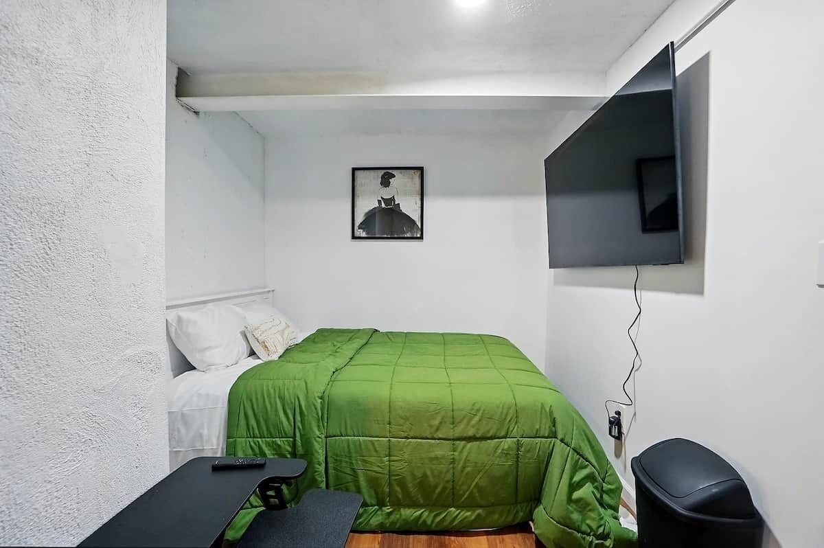 A small, sparsely furnished room with a green bed, a single chair, a desk, a TV mounted on the wall, and a framed picture above the bed.