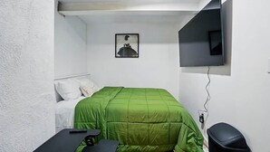 A small, sparsely furnished room with a green bed, a single chair, a desk, a TV mounted on the wall, and a framed picture above the bed. - Modern Studio W/Parking/Long-Term Ready (Cincinnati)