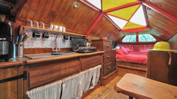 A compact kitchen with wooden cabinets, a stove, and a sink. There is a small dining area with a bench and a red bed in the corner. - Dashing Dennis - 22 (Launceston)