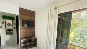 A modern living room with a flat-screen TV mounted on a wooden panel, a sliding glass door leading to a balcony, and a tiled floor.