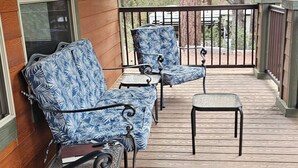 Un porche cubierto con suelo de madera y barandillas de metal. Hay tres sillas con tapicería floral azul y una mesita.