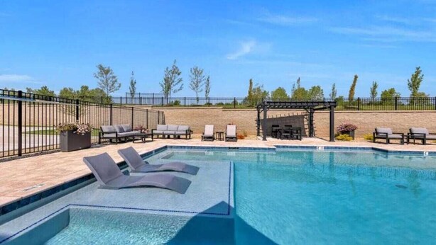 Una piscina rectangular con sillas de descanso y una zona de estar cubierta, rodeada por una valla y árboles.