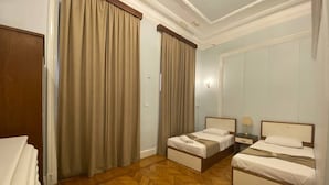 Une chambre d’hôtel avec deux lits simples, un parquet en bois disposé en point de Hongrie et des rideaux de couleur beige.