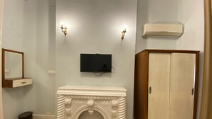 Une pièce avec une cheminée blanche, un téléviseur fixé au mur, deux appliques murales, une armoire et une petite poubelle.