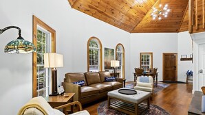 Un salón espacioso con techo de madera, un sofá de cuero marrón, un sillón blanco y un comedor con mesa y sillas de madera.