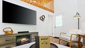 Una sala de estar moderna con chimenea, televisión, una cesta de mimbre, un sillón de mimbre y un letrero de bienvenida sobre una bandeja.