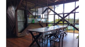 A dining area with a long table and benches, large windows, and a view of greenery. - Coorg Tree House Tent (Madikeri)