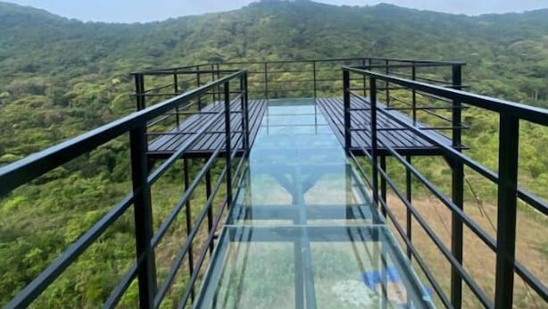Uma ponte de vidro com grades de metal, rodeada por vegetação exuberante e colinas.