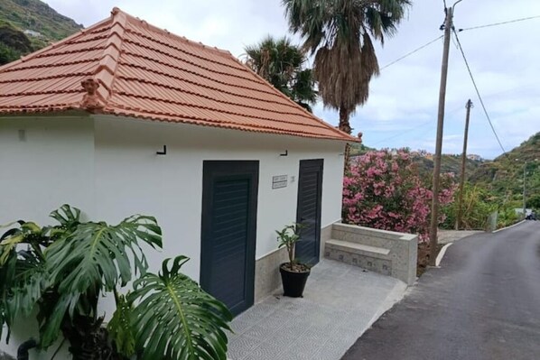 A small, single-story building with a red-tiled roof and a white facade. There are two doors, one of which is closed, and a small potted plant near the entrance. - Traditional Madeiran farm house (Ribeira Brava)