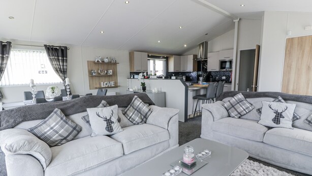 A modern living room with two grey sofas, a coffee table, and a kitchen in the background. - Manor Heights Lodge (Wemyss Bay)