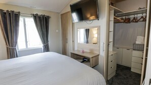 A bedroom with a large bed, a TV mounted above a wooden dresser, and a walk-in closet with hanging clothes. - Manor Heights Lodge (Wemyss Bay)