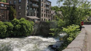 A stone building with multiple windows and balconies, a river with a bridge, and a stop sign on the sidewalk. - Old Post Office Cottage (Ulverston)
