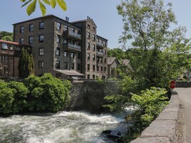 Un edificio de piedra con varias ventanas y balcones, un río con un puente, y una señal de stop en la acera.